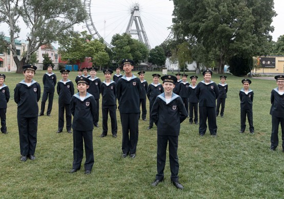 The Vienna Boys Choir standing outdoors at a park with a Ferris Wheel in the background